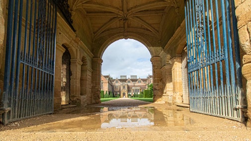 A view of the house taken from the ground, under the Gatehouse arch next to a puddle, with blue skies above.